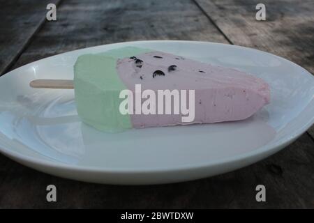 Pink ice cream popsicle in the shape of watermelon on a white plate. Vintage wooden table background with copy space. Summer sweets and desserts. Stock Photo