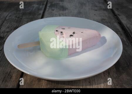 Pink ice cream popsicle in the shape of watermelon on a white plate. Vintage wooden table background with copy space. Summer sweets and desserts. Stock Photo