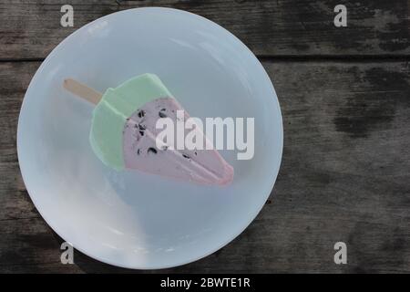 Pink ice cream popsicle in the shape of watermelon on a white plate. Vintage wooden table background with copy space. Summer sweets and desserts. Stock Photo