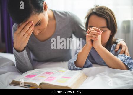 Mother and her daughter reading from bible and praying in their knees near the bed Stock Photo