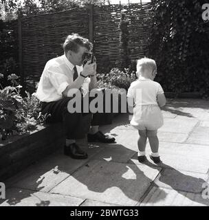 1960s, historical, Outside on a patio, a father sitting on the edge of a flower bed using a lightweight cine camera to film his infant son, England, UK. Small hand-held 8mm cine film cameras were popular in this era for recording moving images of peoples everyday lives, producing amateur films known as 'home movies'. Stock Photo