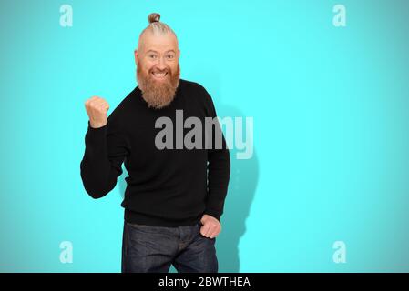 Happy bearded man in black jumper doing winner gesture saying Yes acting like he won something isolated on blue background. Studio portrait. Sincere e Stock Photo