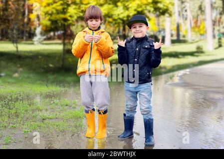 Two cheerfull friendly boys in rubber boots standing in puddle in park showing thumbs up gesture. Concept of social adaptation of autistic children Stock Photo