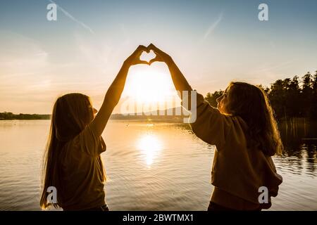 Friends making heart shape with hands against lake during sunset Stock Photo