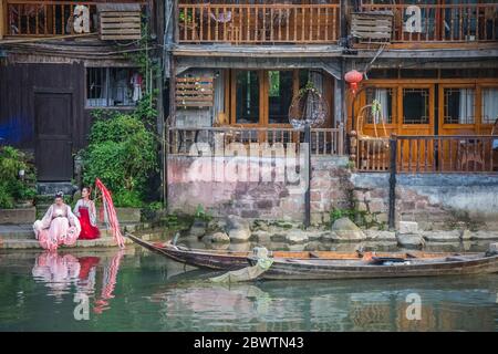Feng Huang, China -  August 2019 : Chinese women dressed in traditional folk costumes sitting and resting on the shore in front of Old historic wooden Stock Photo