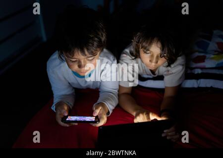 Two brothers lying together on bed at home using electronic devices Stock Photo