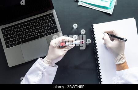 Doctor with COVID-19 sample writing on spiral notebook by laptop at desk in laboratory Stock Photo