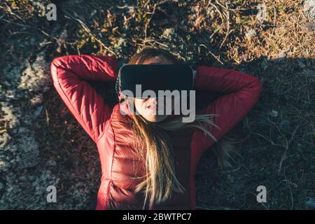 Young woman with VR goggles lying on ground in nature Stock Photo