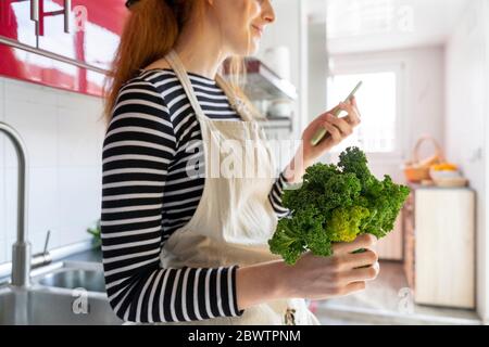 Young woman standing in kitchen, holding cale, using smartphone Stock Photo