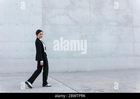 Young woman wearing black suit walking in front of concrete wall Stock Photo
