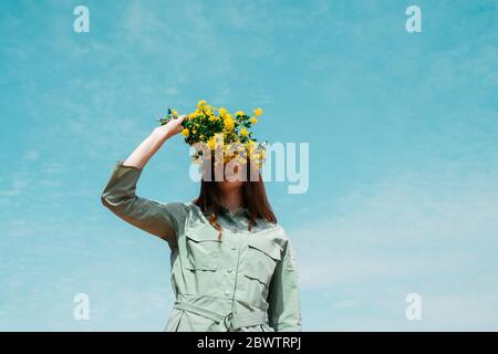 Redheaded young woman against sky covering eyes with bunch of yellow flowers Stock Photo