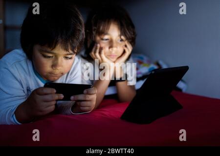 Two brothers lying together on bed at home using electronic devices Stock Photo