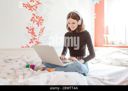 Portrait of teenage girl with headphones sitting on bed using laptop Stock Photo