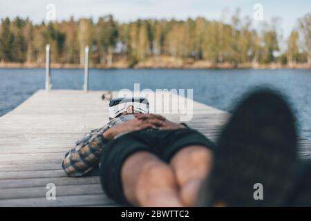 Young man relaxing on jetty, wearing VR glasses Stock Photo