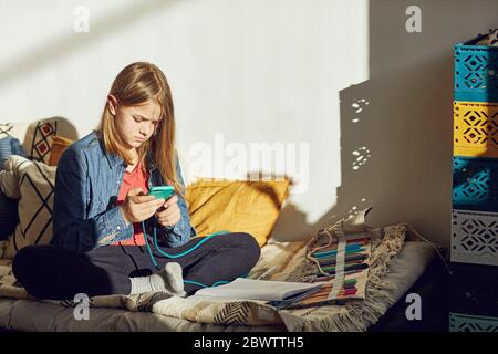 Girl sitting on bed at home doing homework and using smartphone Stock Photo