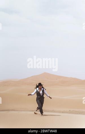 Fashionable young woman dancing barefoot on sand dune, Merzouga desert, Morocco Stock Photo