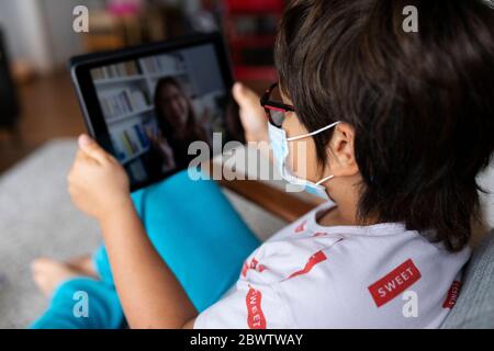 Boy with surgical mask sitting on armchair using digital tablet for video chat with his mother Stock Photo