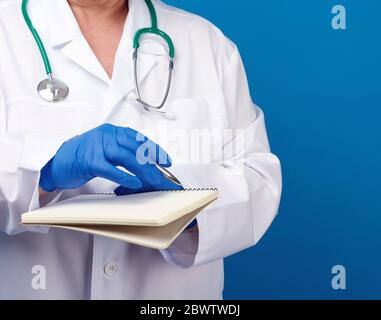 medic woman in white coat, blue latex gloves holding an open notebook with white sheets, blue background, close up Stock Photo