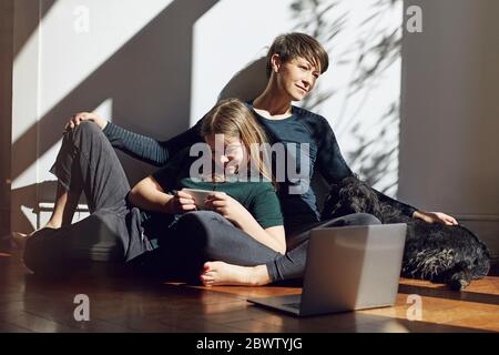 Mother and daughter sitting on the floor at home with dog, laptop and smartphone Stock Photo