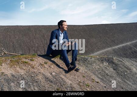 Mature businessman sitting on a disused mine tip holding tablet Stock Photo