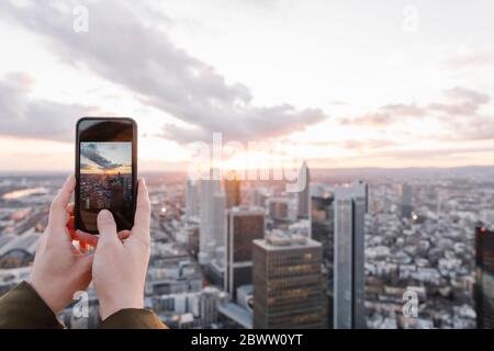 Germany, Hesse, Frankfurt, Hands of woman taking smart phone photos of city downtown at sunset Stock Photo