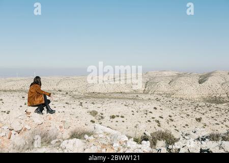 Full length of woman sitting on rocks while looking at desert landscape against clear blue sky Stock Photo