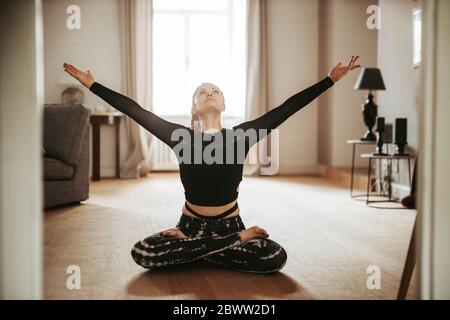 Woman sitting in lotus pose with raised arms at home Stock Photo