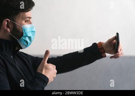 Man wearing mask gesturing thumbs up while taking selfie on smart phone Stock Photo