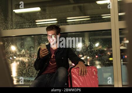 Smiling young man with suitcase sitting at rainy window using smartphone Stock Photo