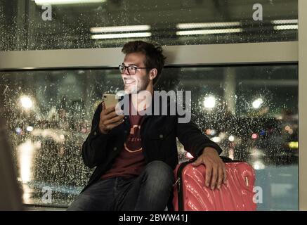 Smiling young man with suitcase sitting at rainy window using smartphone Stock Photo