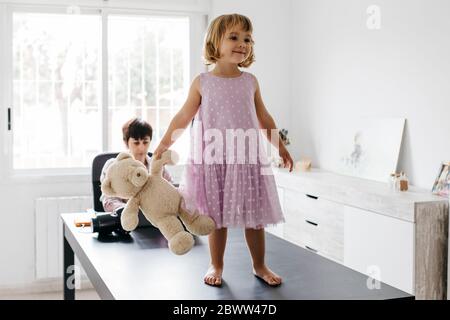 Mother working at home, daughter standing with teddy bear on table Stock Photo