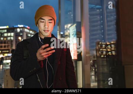 Stylish man with yellow hat and earphones using smartphone at evening on the street at night Stock Photo