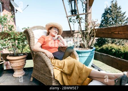 Portrait of pensive mature woman with laptop relaxing on terrace Stock Photo