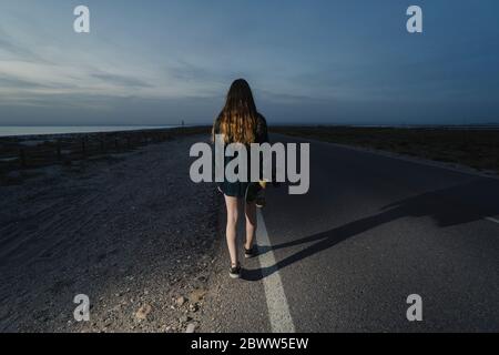 Back view of young woman with skateboard walking along asphalt road, Almeria, Spain Stock Photo