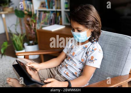 Boy with surgical mask sitting on armchair at home using digital tablet Stock Photo