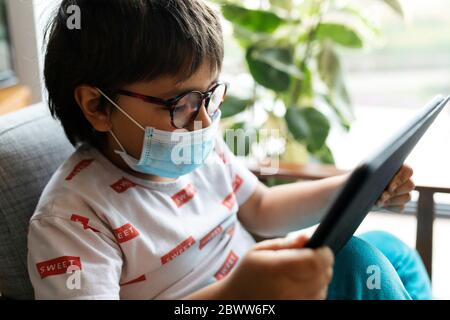 Boy with surgical mask sitting on armchair using digital tablet Stock Photo