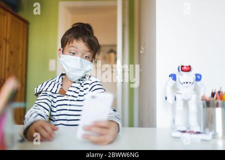 Boy doing homeschooling and using smartphone wearing mask at home during corona crisis Stock Photo
