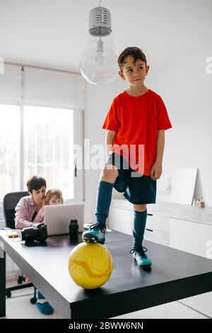 Mom working at home with her children, son standing on soccer ball on table Stock Photo
