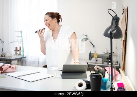 Laughing seamstress working in tailor shop using smartphone and laptop Stock Photo
