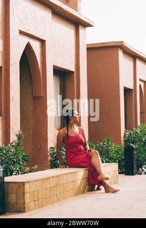 Young woman sitting on wall in front of a house, Merzouga, Morocco Stock Photo