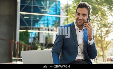 Smiling businessman sitting outside using a cellphone and laptop Stock Photo