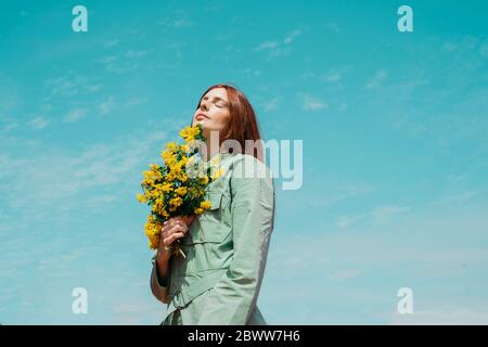 Portrait of redheaded young woman with eyes closed standing against sky holding bunch of yellow flowers Stock Photo