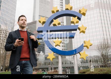 Thoughtful businessman using mobile phone while standing against sculpture and European Central Bank Stock Photo