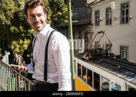 Smiling young man with smartphone and earbuds in the city, Lisbon, Portugal Stock Photo
