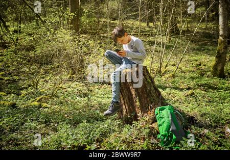 Full length of boy sitting on tree stump while using smart phone in Swabian Jura forest during hiking Stock Photo