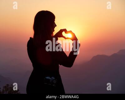 Silhouette of woman shaping heart with her hands at sunset, Serra da Leba, Angola Stock Photo