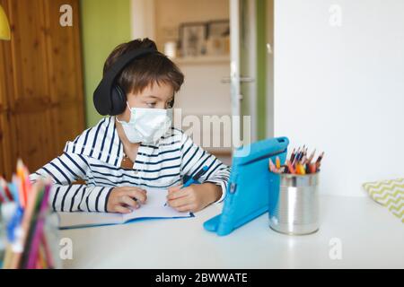 Boy doing homeschooling and writing on notebook, using tablet and headphones, wearing mask at home during corona crisis Stock Photo
