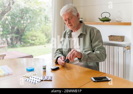 Retired diabetic senior man using glaucometer during blood sugar test while sitting at table Stock Photo