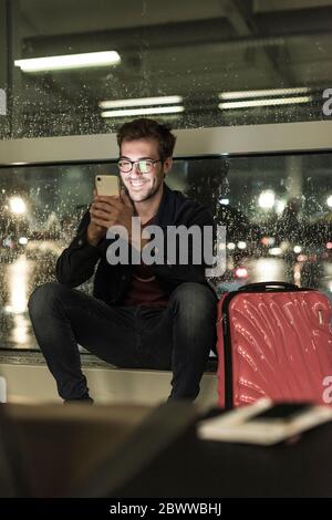 Smiling young man with suitcase sitting at rainy window using smartphone Stock Photo
