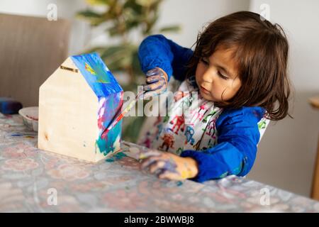 Cute girl painting small wooden model house with paintbrush in living room Stock Photo